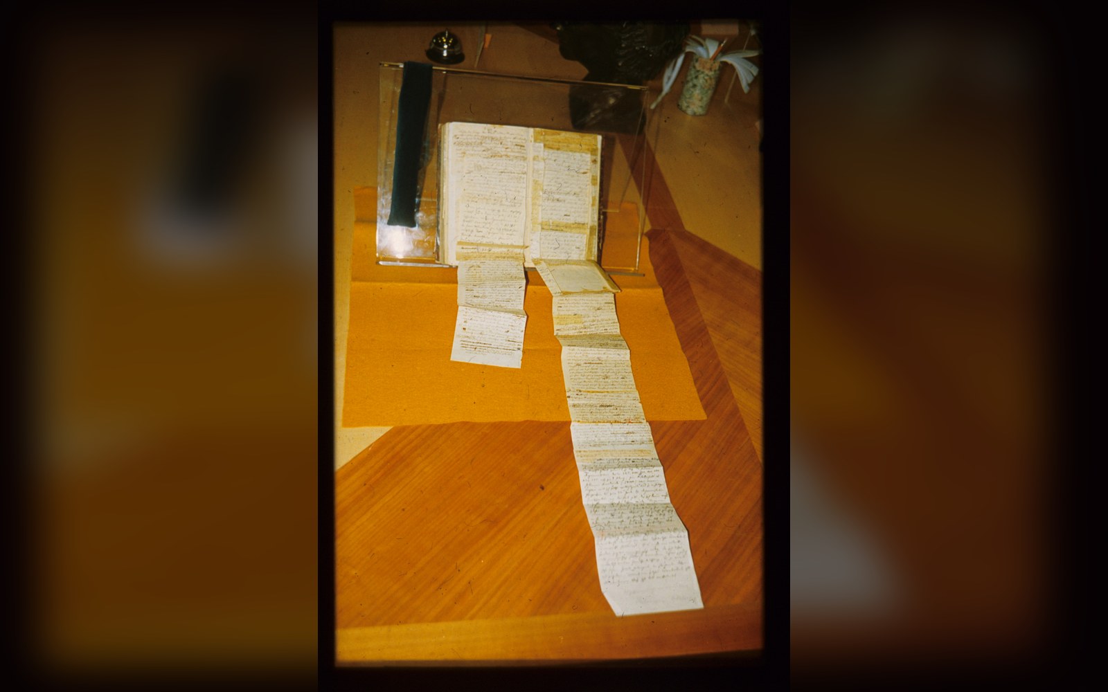 An open handwritten manuscript on a stand, its accordion-folded pages unfolding across a wooden table